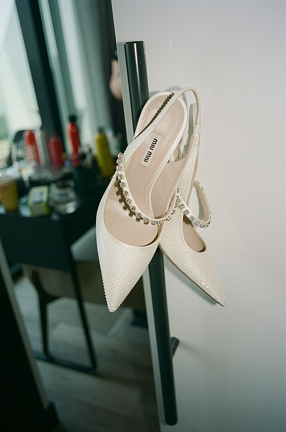 Bridal shoes in white slingback heels with rhinestone ankle straps and beaded texture, resting on a mirror beside hair products in a room