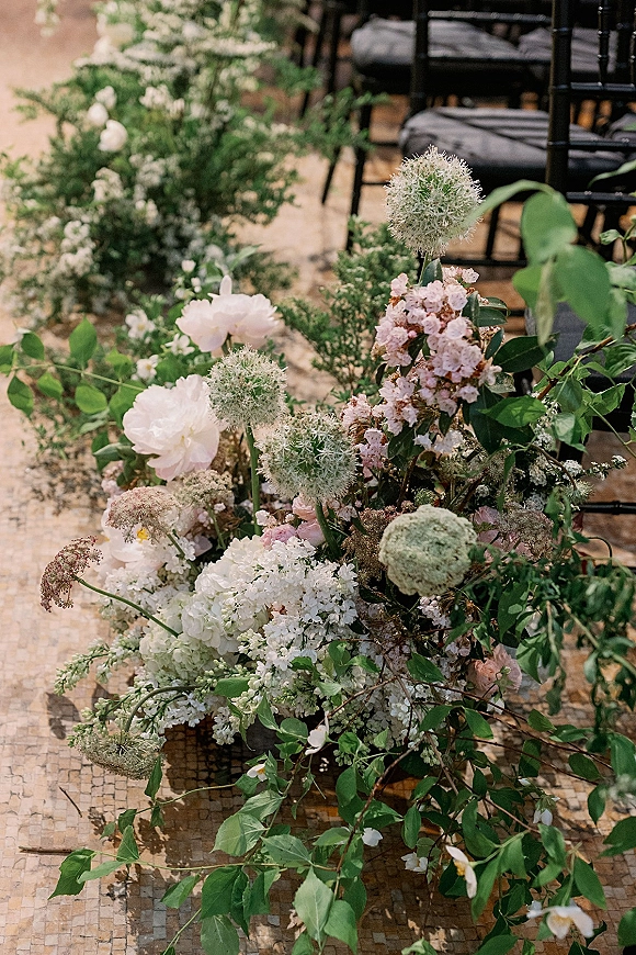Ceremony aisle flowers in white and blush with hydrangea, allium, and greenery lining a stone-tiled aisle beside black chairs