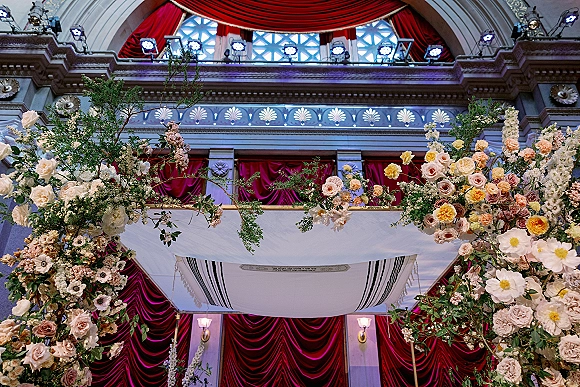 Wedding chuppah decor with a floral chuppah of roses and lush greenery under a canopy in an ornate indoor venue with red drapery