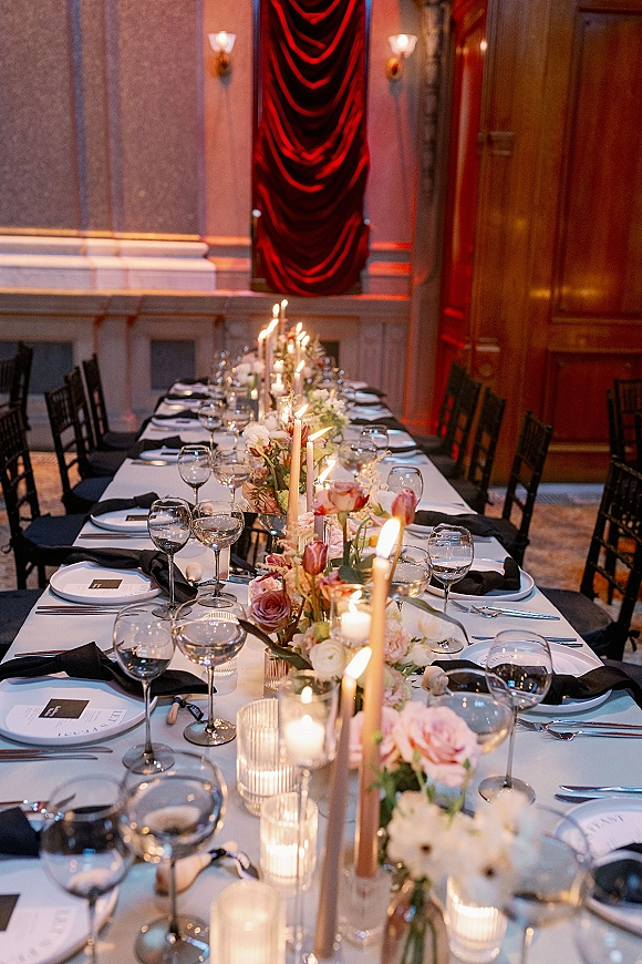 Reception tablescape on a long banquet table wedding with taper candles, floral centerpiece, black napkins, and red velvet drapery backdrop