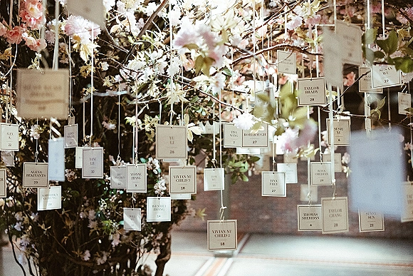 Escort card display with hanging escort cards on strings amid blossoming branches and greenery, arranged against a brick wall indoors