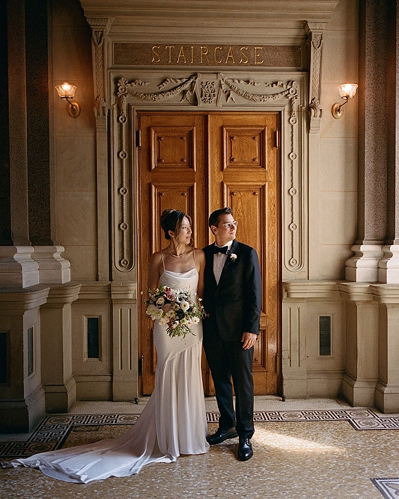 Couple portrait of bride holding a blush and white bouquet beside groom in black tuxedo at an ornate stone doorway with wood doors