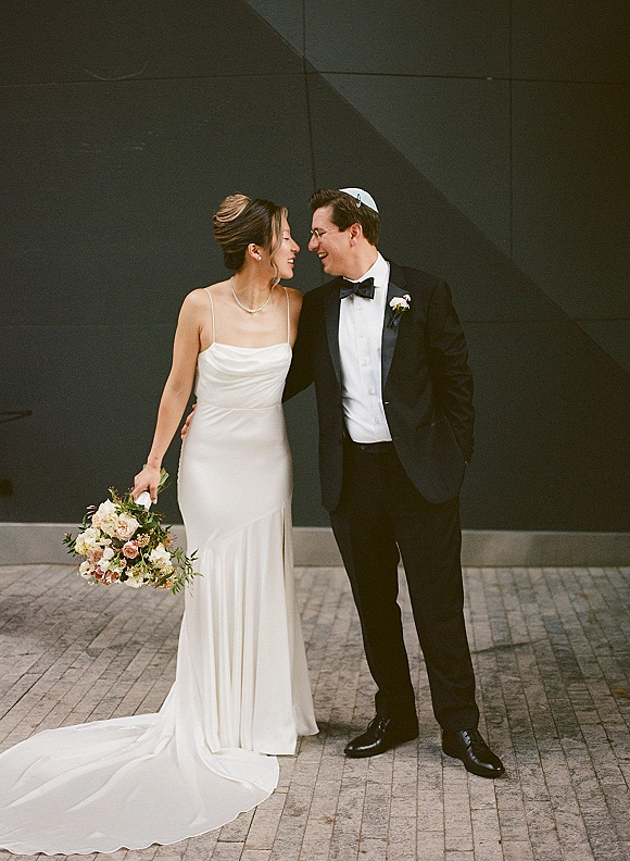 Couple portrait of bride and groom laughing, bride holding bouquet in minimalist gown beside groom in black tuxedo against a dark wall