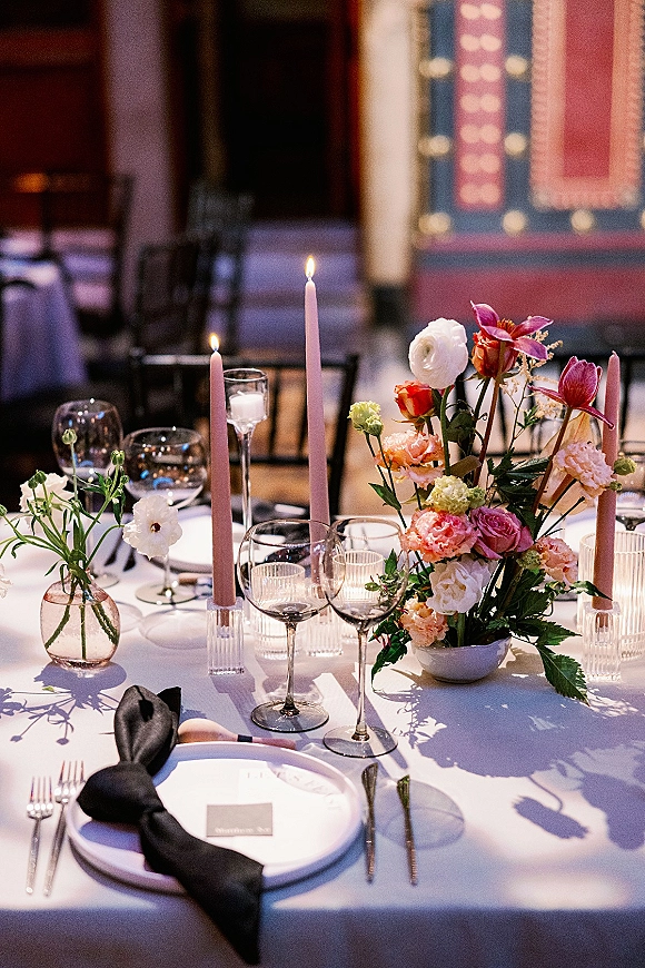 Reception tablescape with wedding table centerpiece of colorful florals, pink taper candles, and black napkin place settings in a dim indoor venue