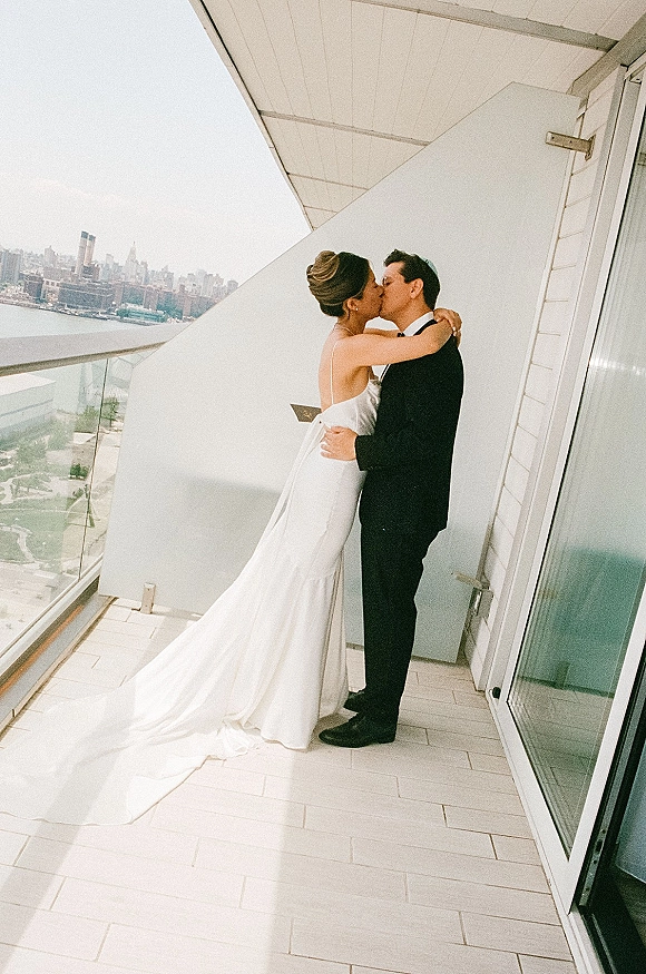 Wedding kiss portrait of bride and groom kissing on a balcony terrace, her long veil and train flowing with city skyline and water behind