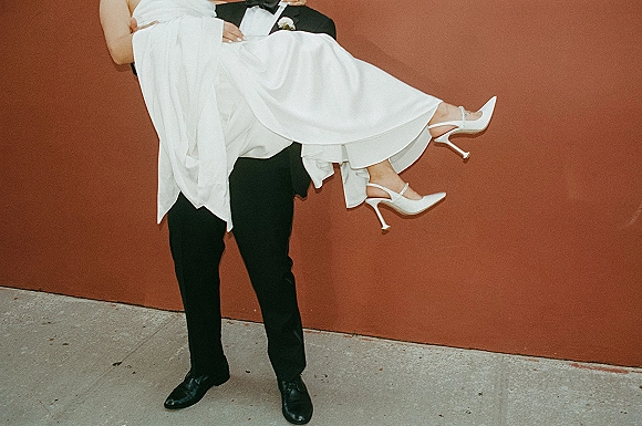 Wedding couple portrait of groom carrying bride, her white slingback heels visible, against a rust wall backdrop on a concrete sidewalk