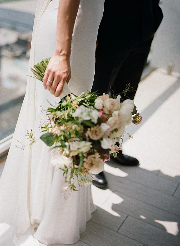 Bridal bouquet of white flowers and greenery in a hand-tied garden style bridal bouquet, held by bride in veil on a sunlit deck