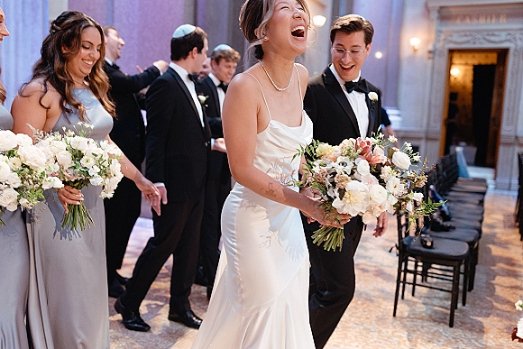 Wedding party moment with bride laughing candid as bridesmaids hold bouquets and groomsmen in tuxedos walk a patterned indoor aisle with sconces