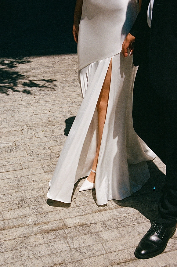 Wedding dress details with a thigh slit wedding dress, showing satin hem, pointed-toe ankle-strap heels, and couple holding hands on stone pavement