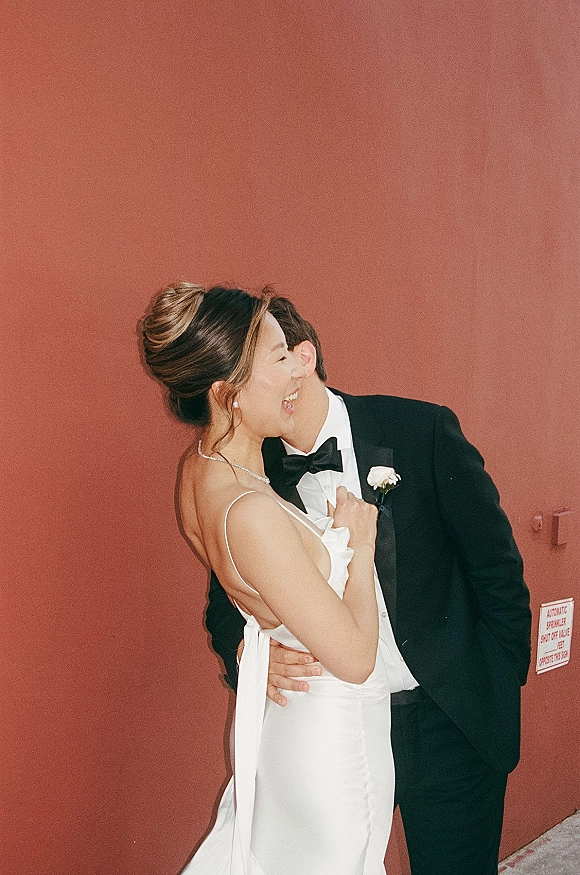 Wedding couple portrait with bride in a satin slip gown and pearl necklace embracing tuxedoed groom against a terracotta wall