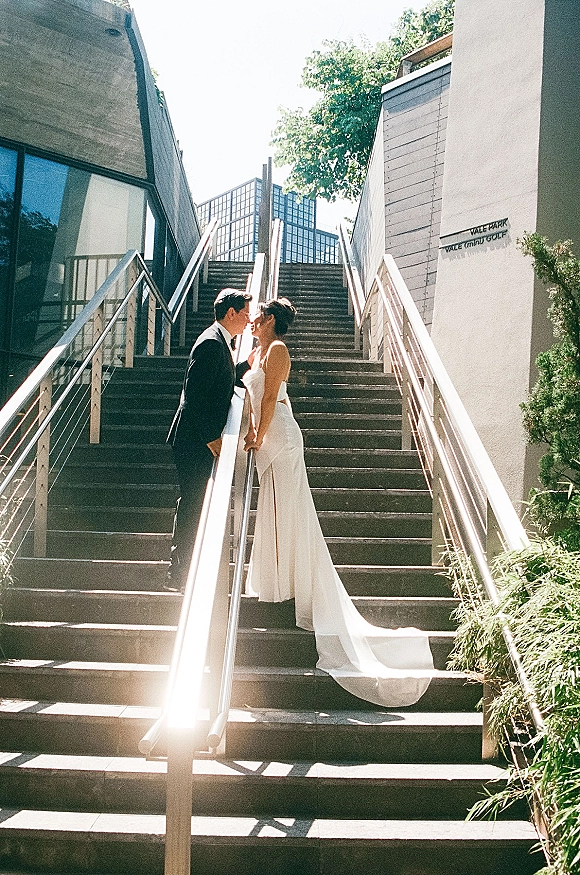 Wedding kiss portrait of bride and groom kissing on an outdoor staircase, her long-train dress flowing beside his tuxedo by modern windows