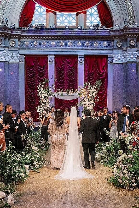 Wedding processional as bride walking down aisle from behind, cathedral veil trailing in a grand hall with red velvet drapes and guests