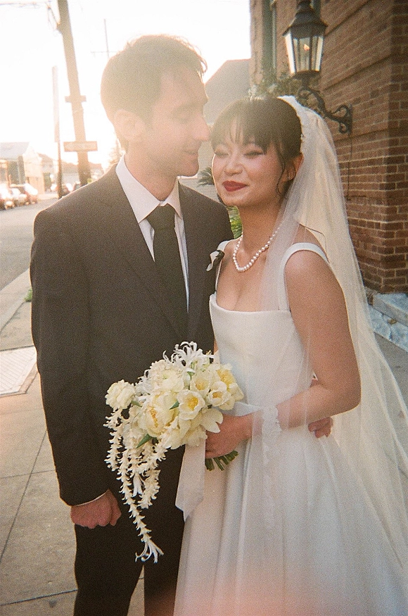 Couple portrait of bride and groom hugging on a sunlit city street, her veil and pearl necklace glowing as she holds a bouquet