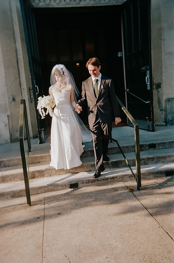 Wedding recessional as bride and groom walking hand in hand down stone steps, her long veil trailing and bouquet held near open doors