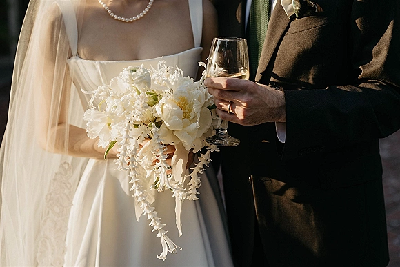 Wedding couple portrait with bride and groom close up, holding champagne flutes as bride shows bouquet and rings in sunlit greenery