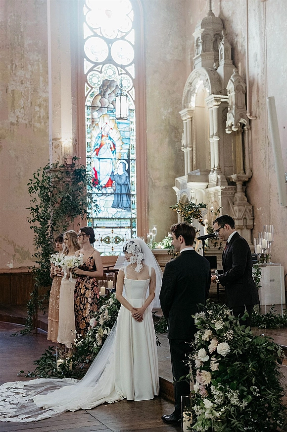 Wedding ceremony as bride and groom stand at the church altar with officiant, lace veil, bouquets, candles, and stained glass backdrop