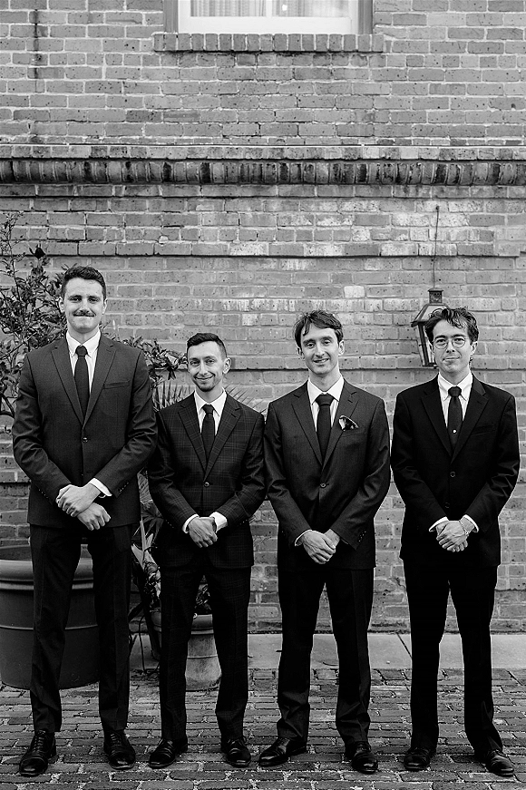 Groomsmen portrait of men in matching suits and ties, one wearing eyeglasses, posed by a brick wall with plants and lanterns