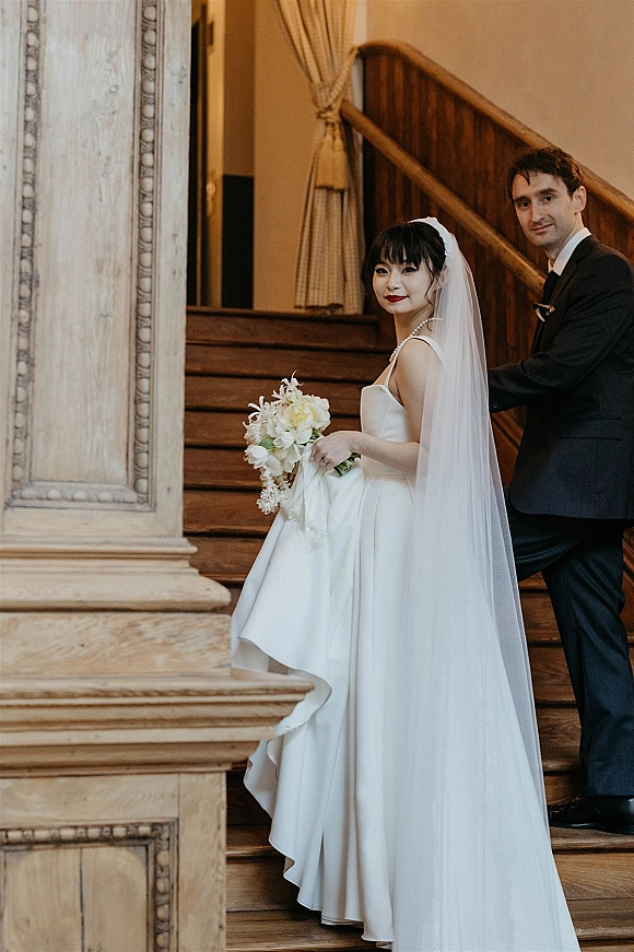 Couple portrait of bride and groom on stairs, bride in strapless satin dress with long veil holding a cascading white orchid bouquet