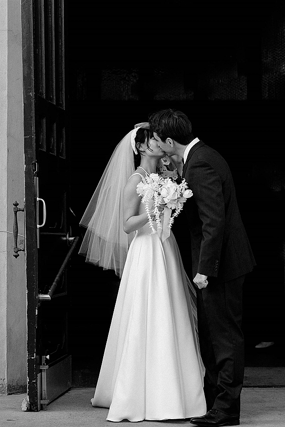 Wedding kiss portrait of bride and groom kissing in a doorway, bride in veil with bouquet, groom in dark suit against dark interior
