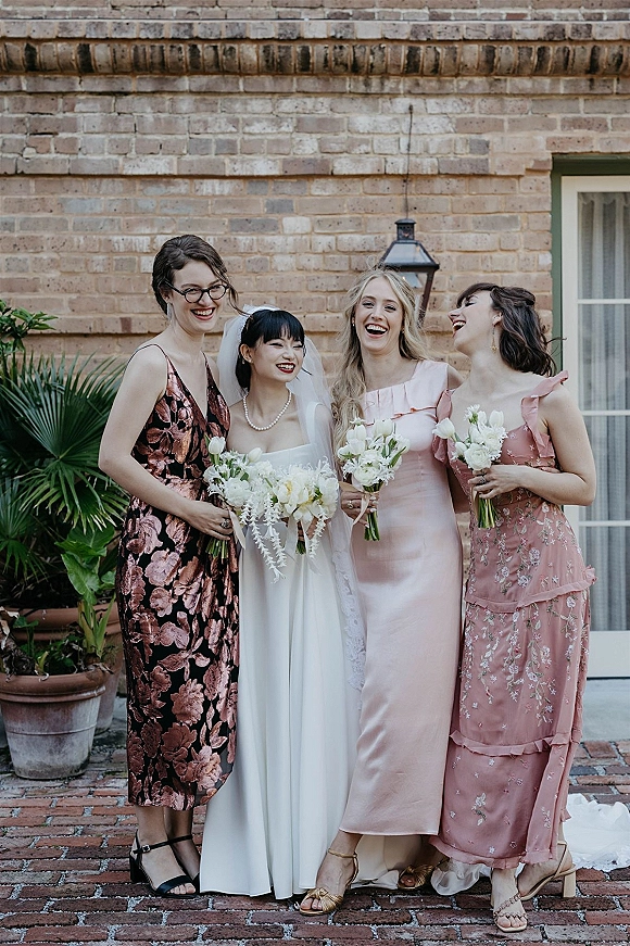 Bride with bridesmaids in pink dresses holding white bouquets, her veil draped over a pearl necklace against a brick courtyard wall