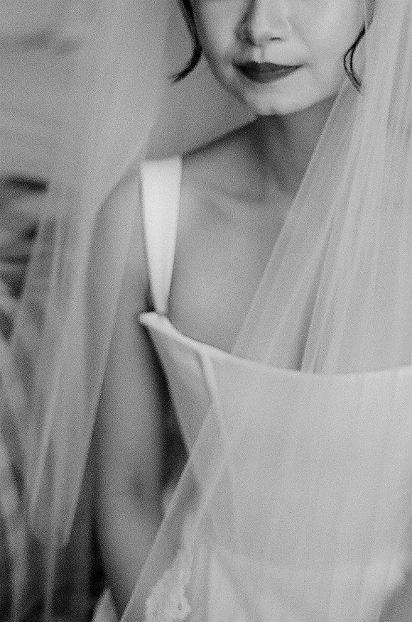 Bride portrait in black and white with a bridal veil draped over her shoulder, spaghetti strap dress and lipstick, gazing away by a plain wall