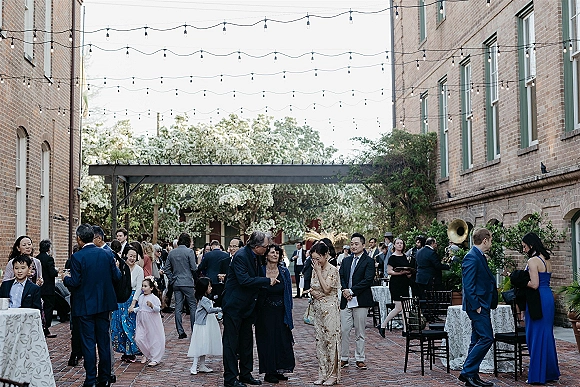Cocktail hour with wedding guests mingling around cocktail tables under string lights in a brick courtyard, holding drinks in formal attire