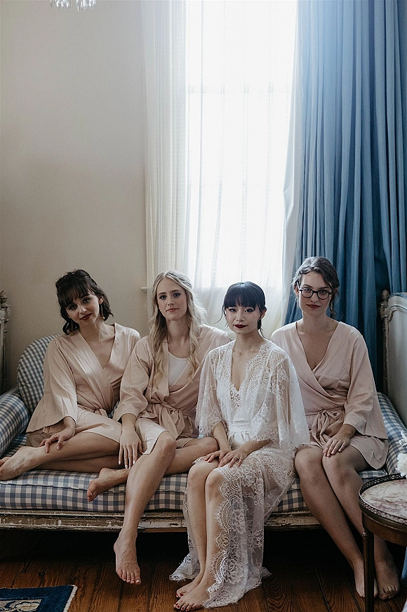 Bride with bridesmaids in blush satin robes and a lace robe gathered by a window with sheer curtains and blue drapes in a bridal suite