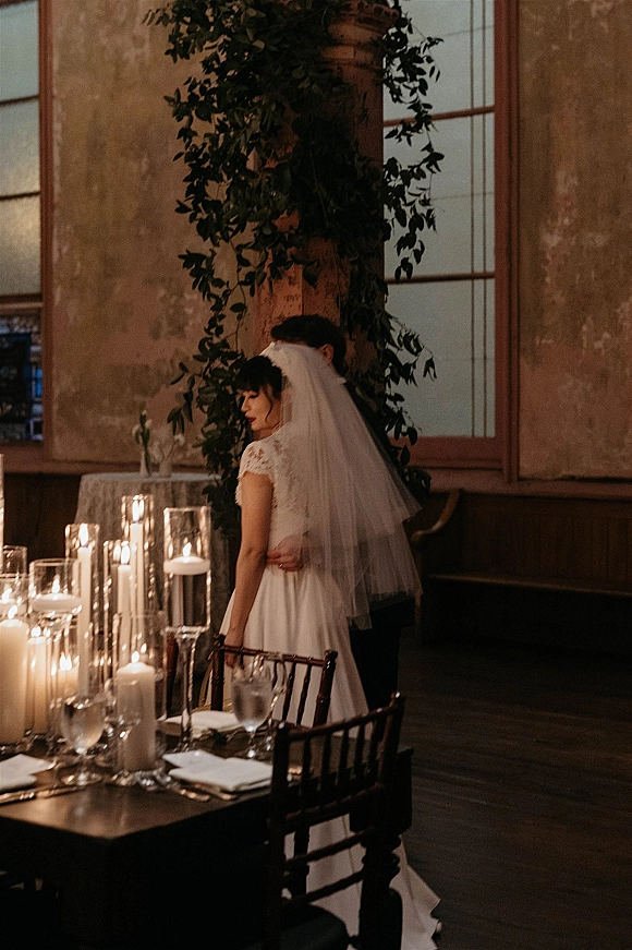 Couple portrait of bride and groom embrace by a candlelit reception table, her veil and lace sleeves glowing in a rustic loft interior