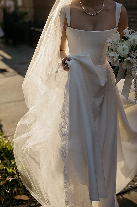 Wedding dress with square neckline, paired with a bridal veil and pearl necklace, as bride holds a white bouquet on a sunlit sidewalk
