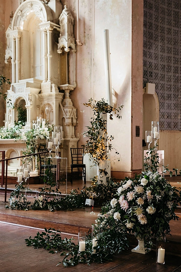 Ceremony altar decor with church wedding altar flowers, greenery garland, pillar candles in glass cylinders, and white urns on clear pedestals