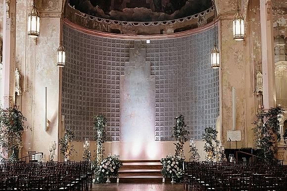 Ceremony setup with indoor wedding ceremony aisle candles, dark wood chairs, and white floral greenery at an ornate church altar with lanterns