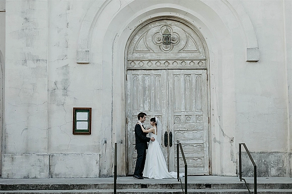 Couple portrait of bride and groom embrace, forehead touching at arched weathered church doors, her veil draped over stone steps