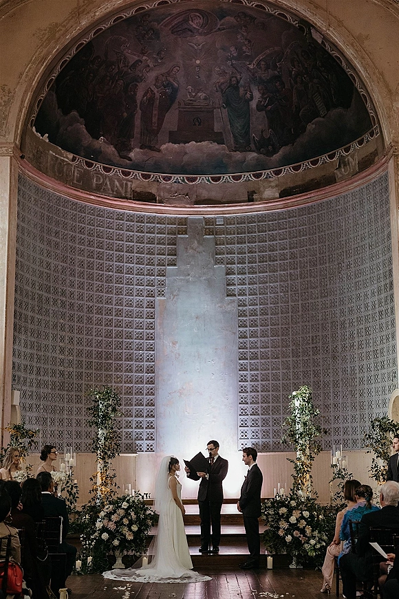 Wedding ceremony with bride and groom exchanging vows as officiant reads, framed by white floral pillars in a mural-ceiling church interior