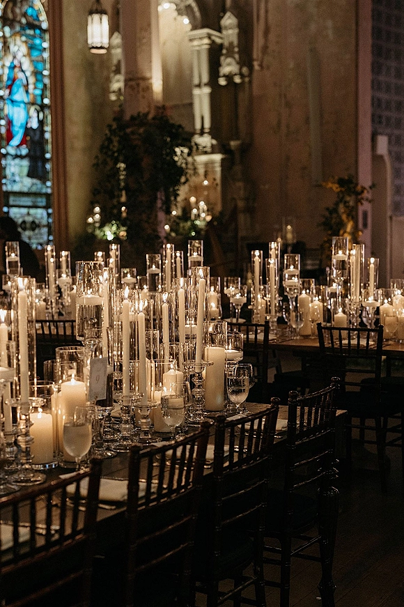 Candlelit reception tablescape with wedding candle centerpiece of taper and pillar candles in glass hurricanes, set in a dim church with stained glass