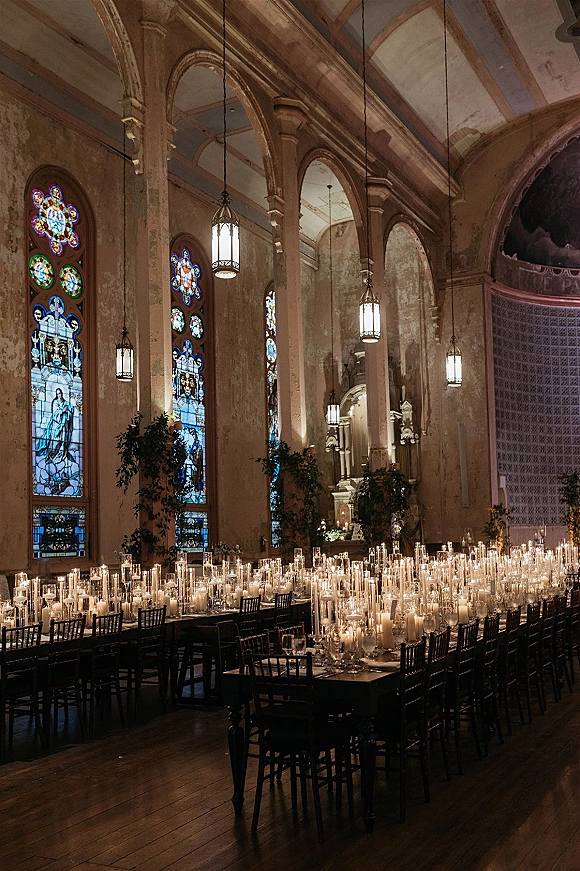 Reception tablescape with candlelit wedding reception taper candles, white place settings and greenery on long tables in a stained-glass church interior