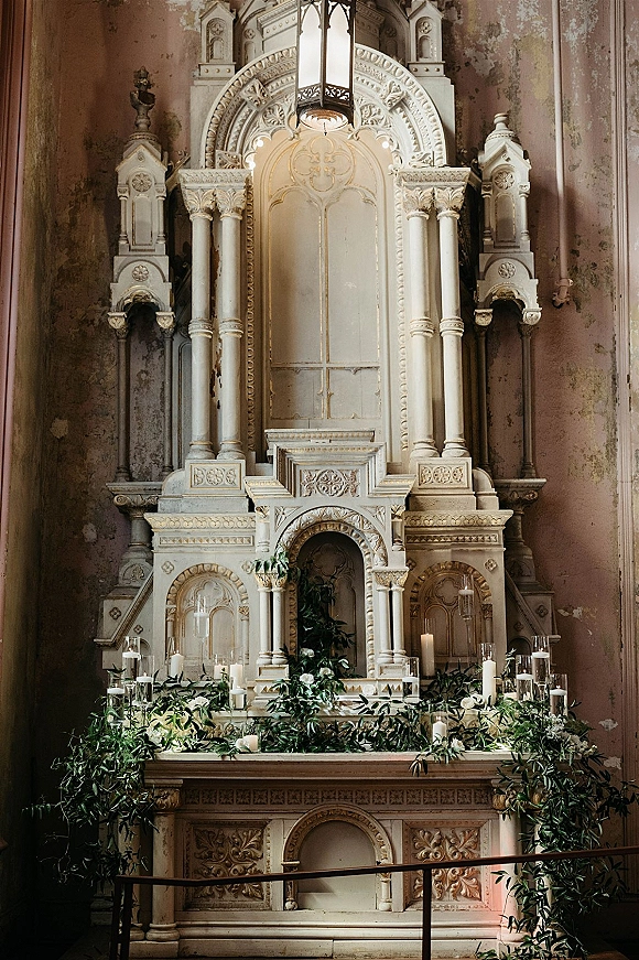 Wedding altar decor with pillar candles in hurricane glass holders, greenery garland and floral arrangement on an ornate church backdrop