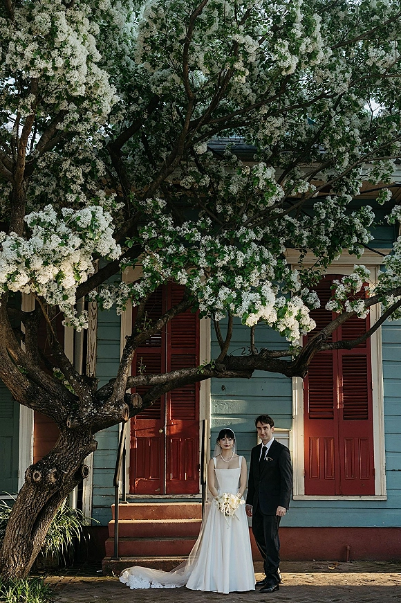 Couple portrait of bride in veil holding a bouquet beside groom in black suit under a flowering tree by blue house with red shutters