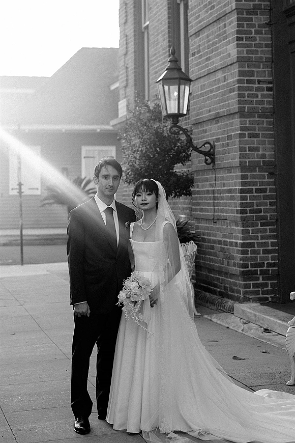 Wedding couple portrait in black and white wedding portrait style, bride in long veil with bouquet beside groom in suit by brick building lantern