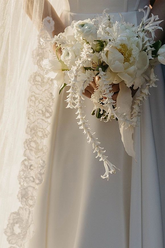 Bridal bouquet of white peonies and ranunculus cascading with a ribbon, held against a bridal gown and veil in soft indoor light