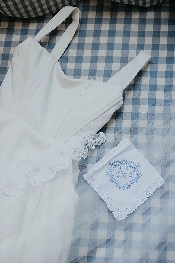 Wedding dress details in a bridal dress flat lay with lace trim bodice, tulle veil, and embroidered handkerchief on blue gingham bedding