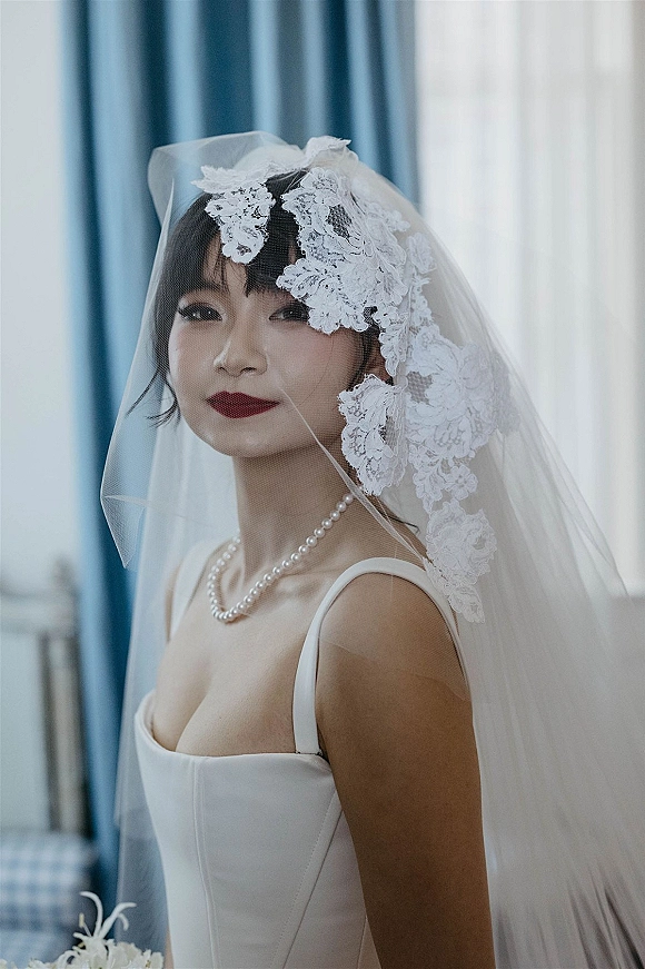 Bridal portrait with lace mantilla veil over her face, pearl necklace and red lipstick, lit by window light against blue curtains indoors