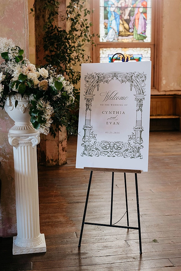 Wedding welcome sign with calligraphy on an easel beside a white floral urn arrangement, set on wood floors near a stained glass window