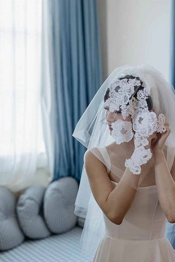 Bridal portrait with lace wedding veil draped over her face, wearing a gown with sheer straps in soft window light by blue curtains