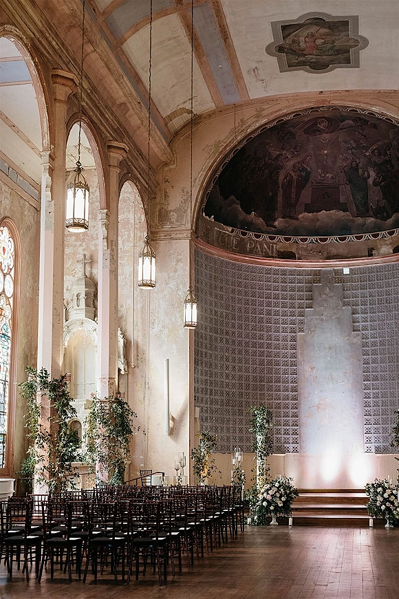 Church ceremony setup with rows of chairs, greenery garlands, and floral urns lining a candlelit aisle beneath stained glass windows