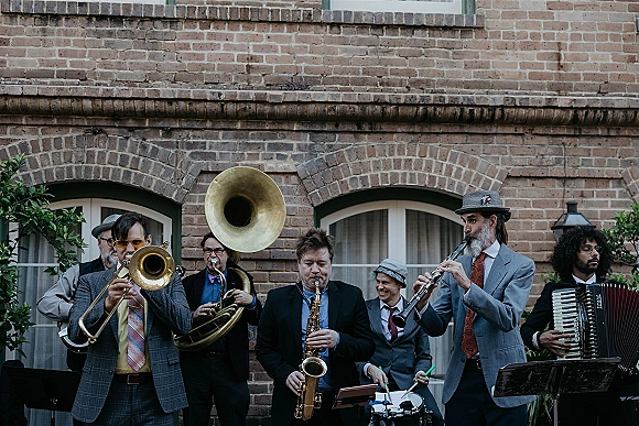 Wedding band playing live wedding music with brass instruments in suits and hats, set in an outdoor courtyard by a brick building with arched windows