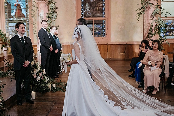 Ceremony moment as bride walking down aisle in classic white bridal gown with long veil, bouquet, and candles in stained glass room