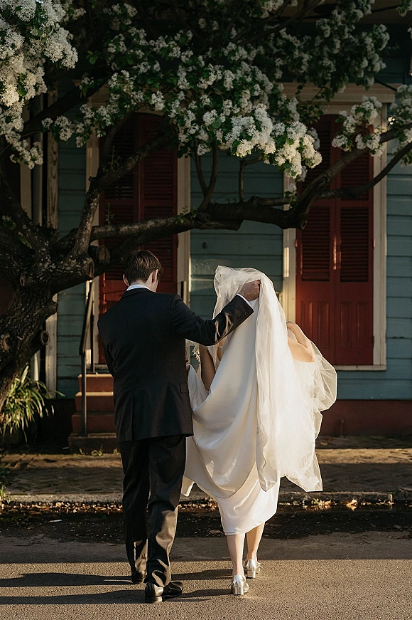 Couple portrait of bride and groom walking away hand in hand, groom lifting her veil by a flowering tree on a quiet street
