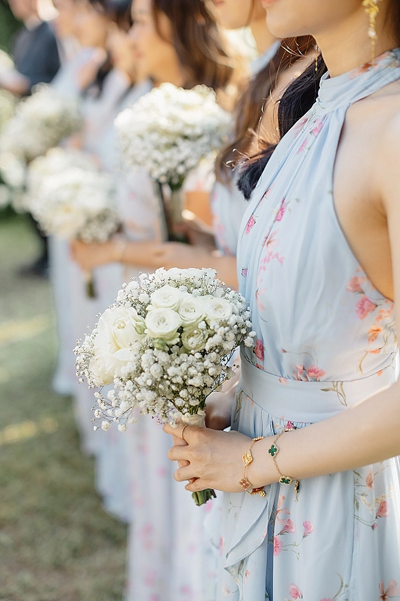 Bridesmaid portrait holding bouquet of white roses and baby's breath, wearing a light blue floral dress and gold earrings on an outdoor lawn