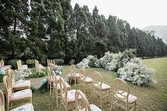 Outdoor ceremony setup with garden wedding ceremony seating, wood chairs and programs flanking a floral lined aisle on a lawn beneath evergreens