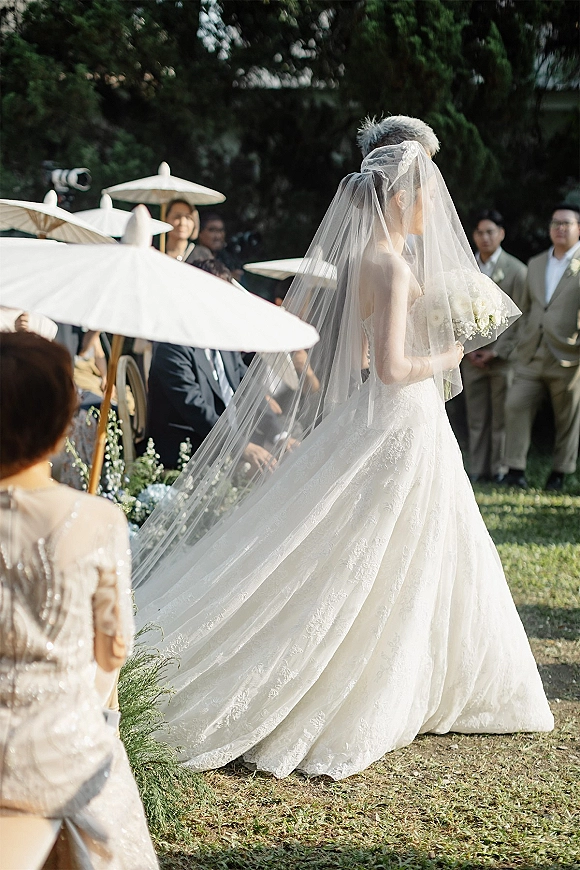 Bridal processional as the bride walking down aisle in a lace gown with long veil and bouquet past guests under white parasols in a sunny garden